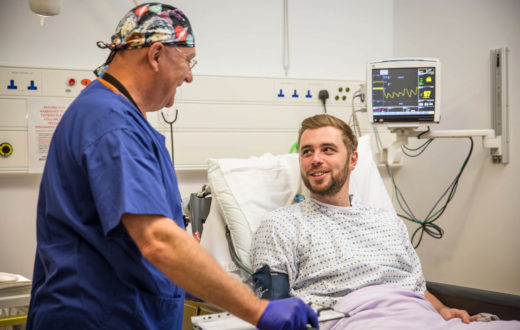 A smiling surgeon in blue scrubs talks to a male patient in a hospital bed with a heart monitor in the background.