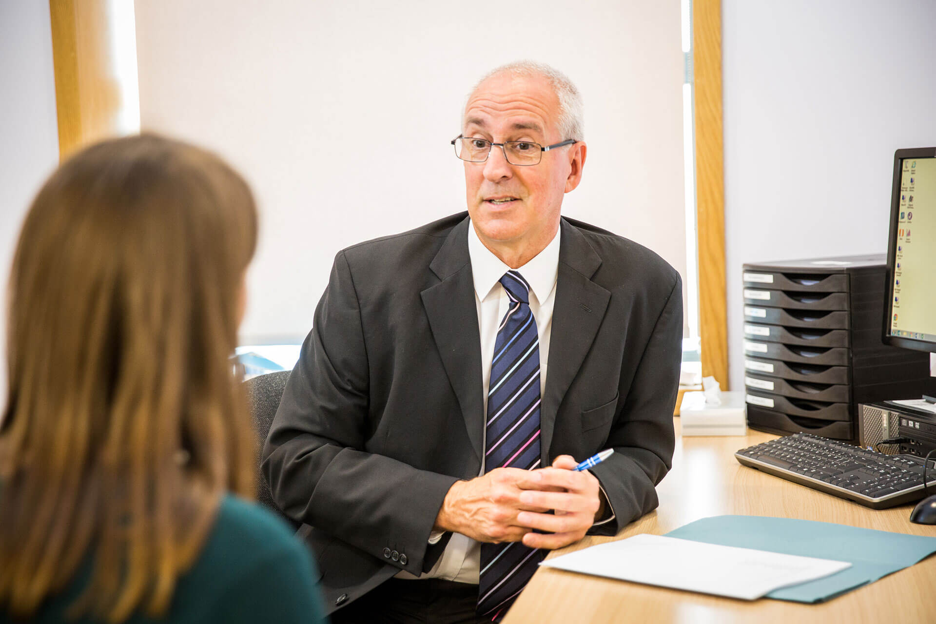 A male consultant in a suit and glasses speaks with a patient during a consultation at KIMS Hospital.