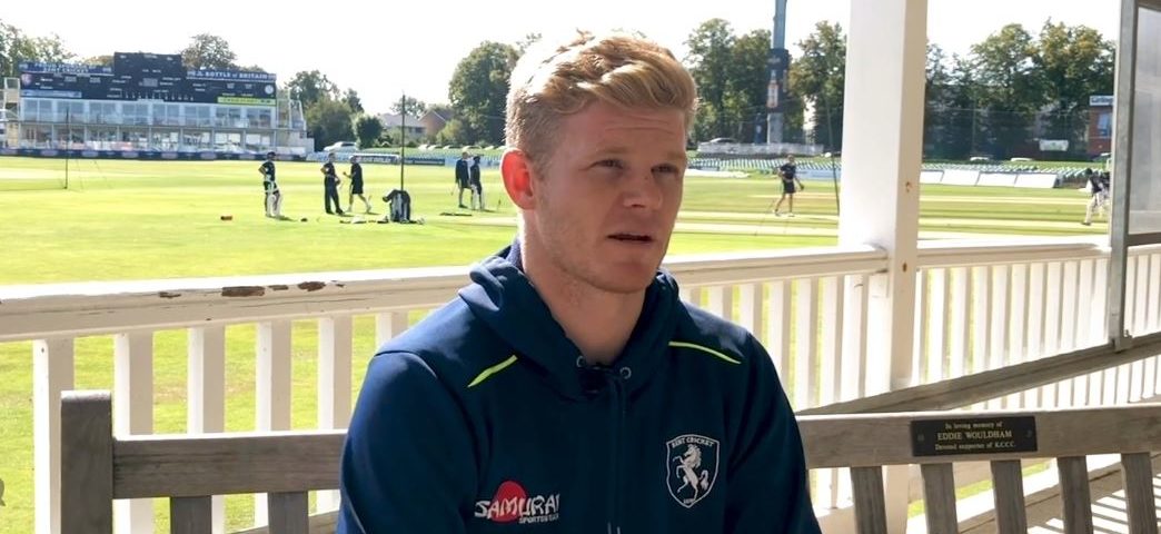 A blonde cricketer in a navy Samurai hoodie sits on a bench during an interview at a sunny Kent cricket ground.