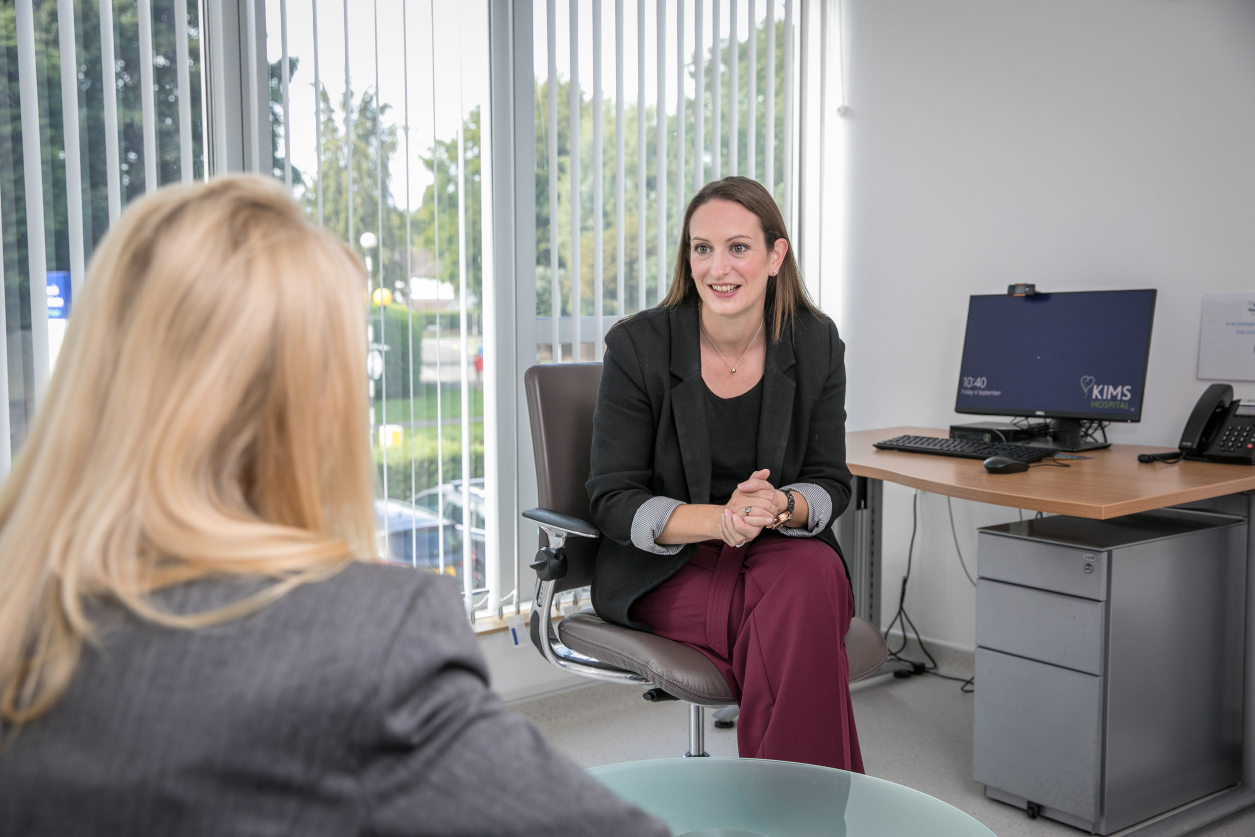 A female professional in a black blazer and maroon trousers speaks with a colleague in a bright KIMS Hospital office.