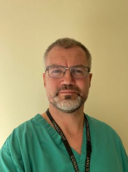 A professional headshot of a man with glasses and a beard wearing green medical scrubs against a plain yellow wall.