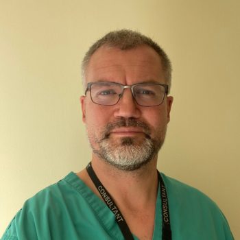 A professional headshot of a man with glasses and a beard wearing green medical scrubs against a plain yellow wall.