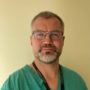 A professional headshot of a man with glasses and a beard wearing green medical scrubs against a plain yellow wall.