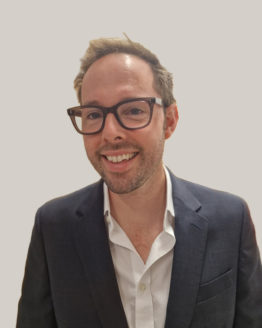 Headshot of a man with dark hair and glasses, wearing a white shirt and dark blazer against a plain grey background.