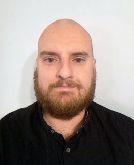 A bald man with a thick brown beard and blue eyes wears a black collared shirt against a plain white background.