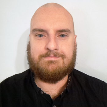 A bald man with a thick brown beard and blue eyes wears a black collared shirt against a plain white background.