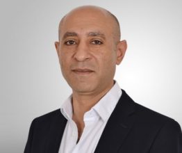 Headshot of a bald man with olive skin wearing a dark suit and white collared shirt against a light grey background.