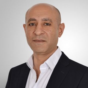 Headshot of a bald man with olive skin wearing a dark suit and white collared shirt against a light grey background.