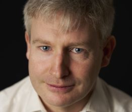 Headshot of a man with short grey hair and fair skin, wearing a white shirt against a dark background.