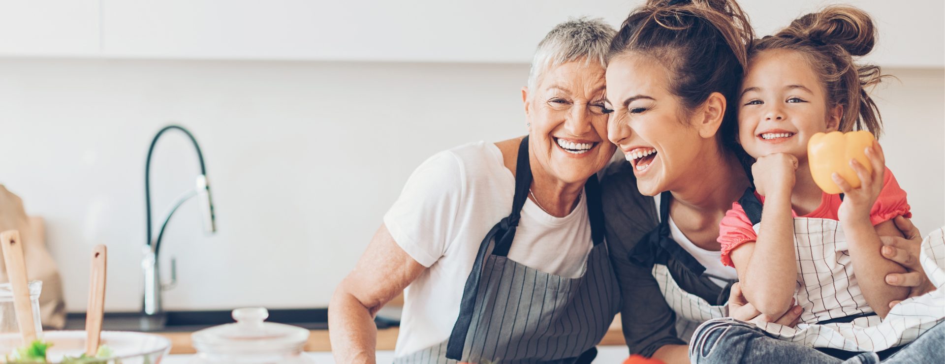 Three generations of women laughing together in a bright kitchen while wearing aprons and holding a yellow pepper.