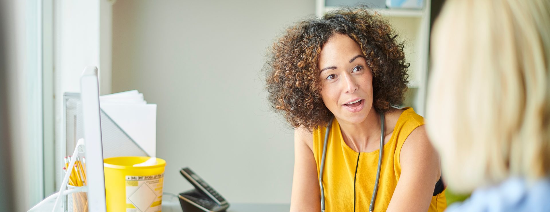 A doctor with curly hair wearing a yellow top and stethoscope speaks with a patient in a bright medical office.