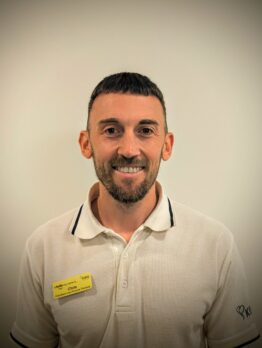 Headshot of Chris Andrews smiling, wearing a white polo shirt with a yellow name badge against a plain background.