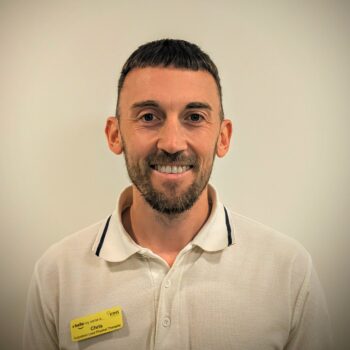 Headshot of Chris Andrews smiling, wearing a white polo shirt with a yellow name badge against a plain background.