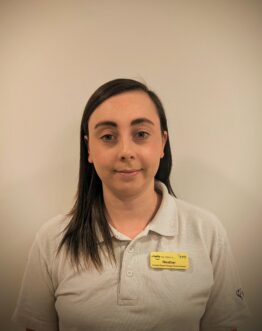 Headshot of a woman with long dark hair, wearing a beige polo shirt and a yellow name tag on a plain background.