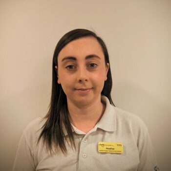 Headshot of a woman with long dark hair, wearing a beige polo shirt and a yellow name tag on a plain background.