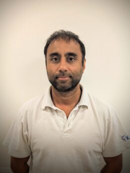 Headshot of a man with short dark hair and a beard, wearing a white polo shirt against a plain light background.