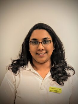 Headshot of a smiling woman with long dark wavy hair and glasses, wearing a white polo shirt on a plain background.
