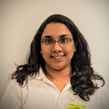 Headshot of a smiling woman with long dark wavy hair and glasses, wearing a white polo shirt on a plain background.