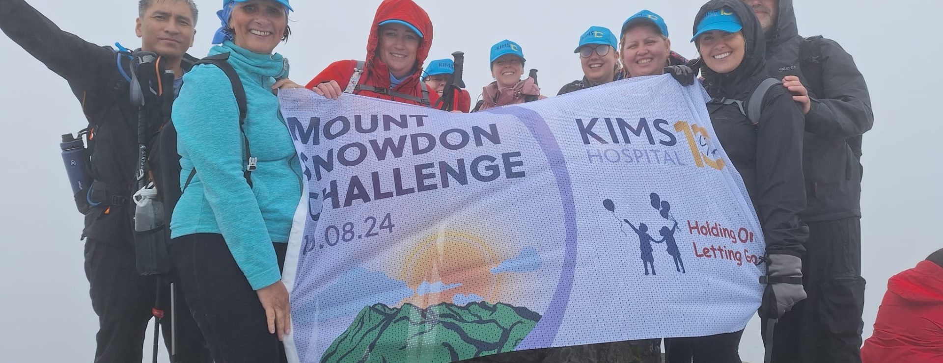 A group of hikers holds a "Mount Snowdon Challenge" banner for KIMS Hospital at the misty summit of a mountain.
