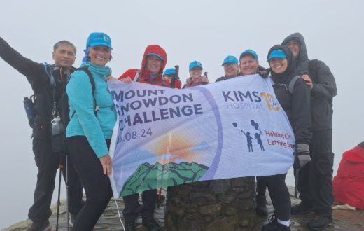 A group of hikers holds a "Mount Snowdon Challenge" banner for KIMS Hospital at the misty summit of a mountain.