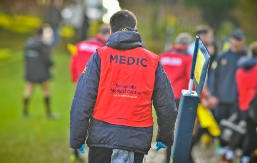 A Sevenoaks Medical Centre medic in a red "MEDIC" vest walks across a rugby pitch to provide medical support.