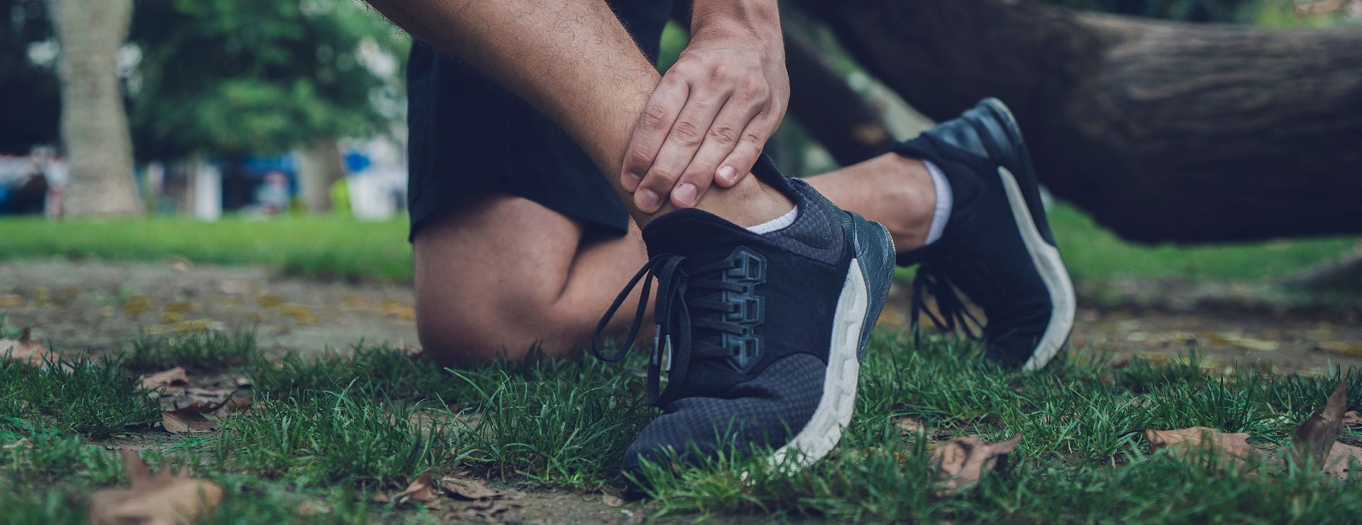 A close-up of a person kneeling on grass while clutching a painful ankle injury during an outdoor workout.