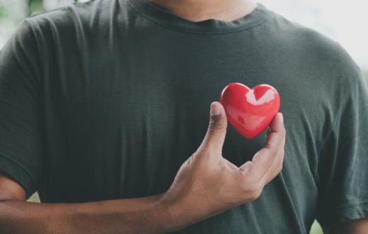 A person in a dark green t-shirt holding a small, glossy red heart against their chest with their right hand.