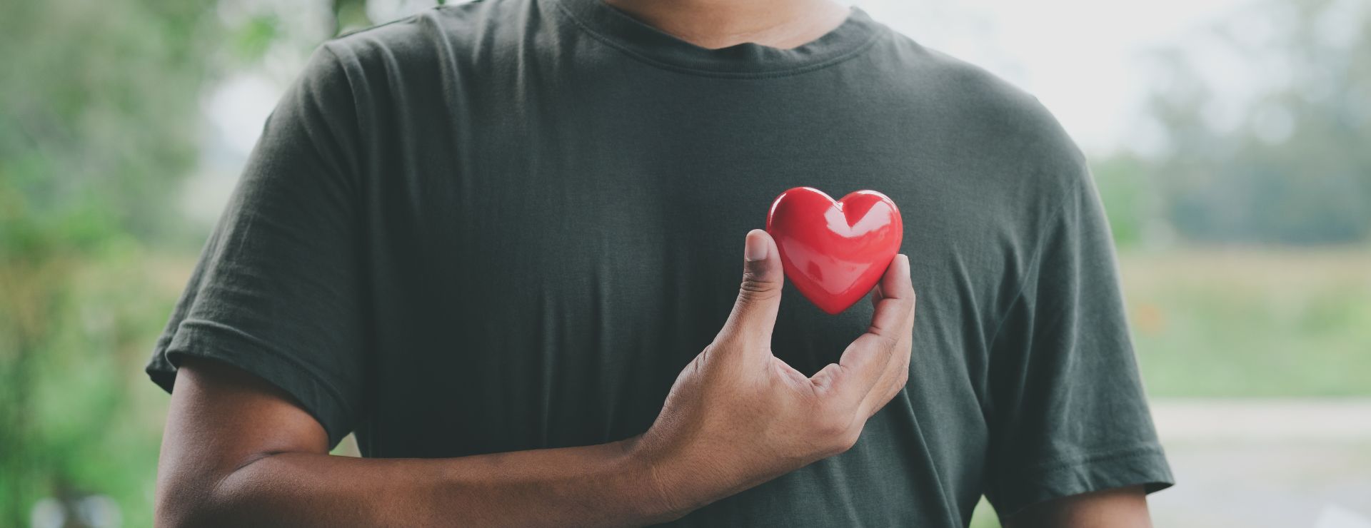 A person in a dark green t-shirt holding a small, glossy red heart against their chest with their right hand.