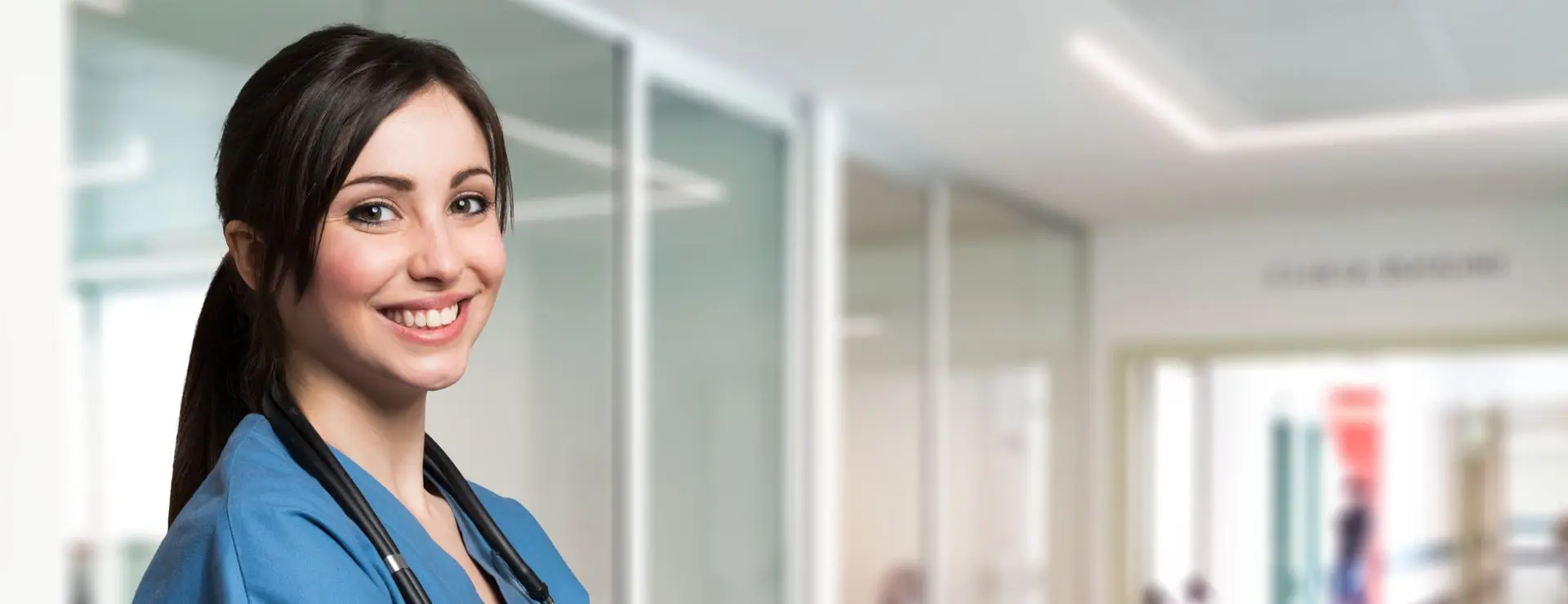 A smiling female healthcare worker in blue scrubs with a stethoscope around her neck, in a bright hospital hallway.