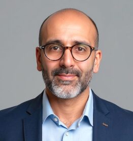 Headshot of a bald man with a grey beard and glasses, wearing a light blue shirt and navy blazer on a grey background.