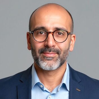 Headshot of a bald man with a grey beard and glasses, wearing a light blue shirt and navy blazer on a grey background.