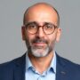 Headshot of a bald man with a grey beard and glasses, wearing a light blue shirt and navy blazer on a grey background.