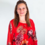 A smiling woman with long brown hair, wearing a red floral patterned top, against a plain light grey background.