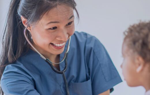A smiling paediatric clinician in blue scrubs using a stethoscope to examine a young patient at KIMS Hospital.