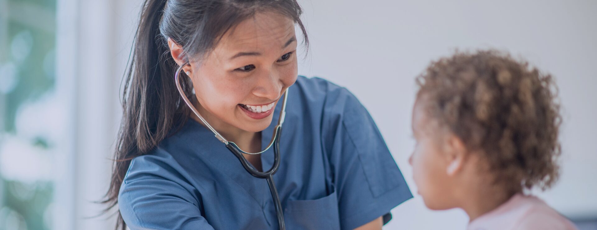 A smiling paediatric clinician in blue scrubs using a stethoscope to examine a young patient at KIMS Hospital.