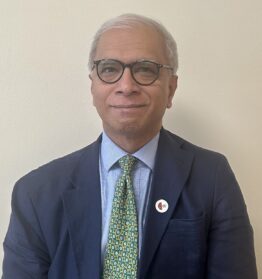 Headshot of Professor Ala wearing glasses, a navy suit, and a patterned green tie against a plain background.
