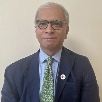 Headshot of Professor Ala wearing glasses, a navy suit, and a patterned green tie against a plain background.