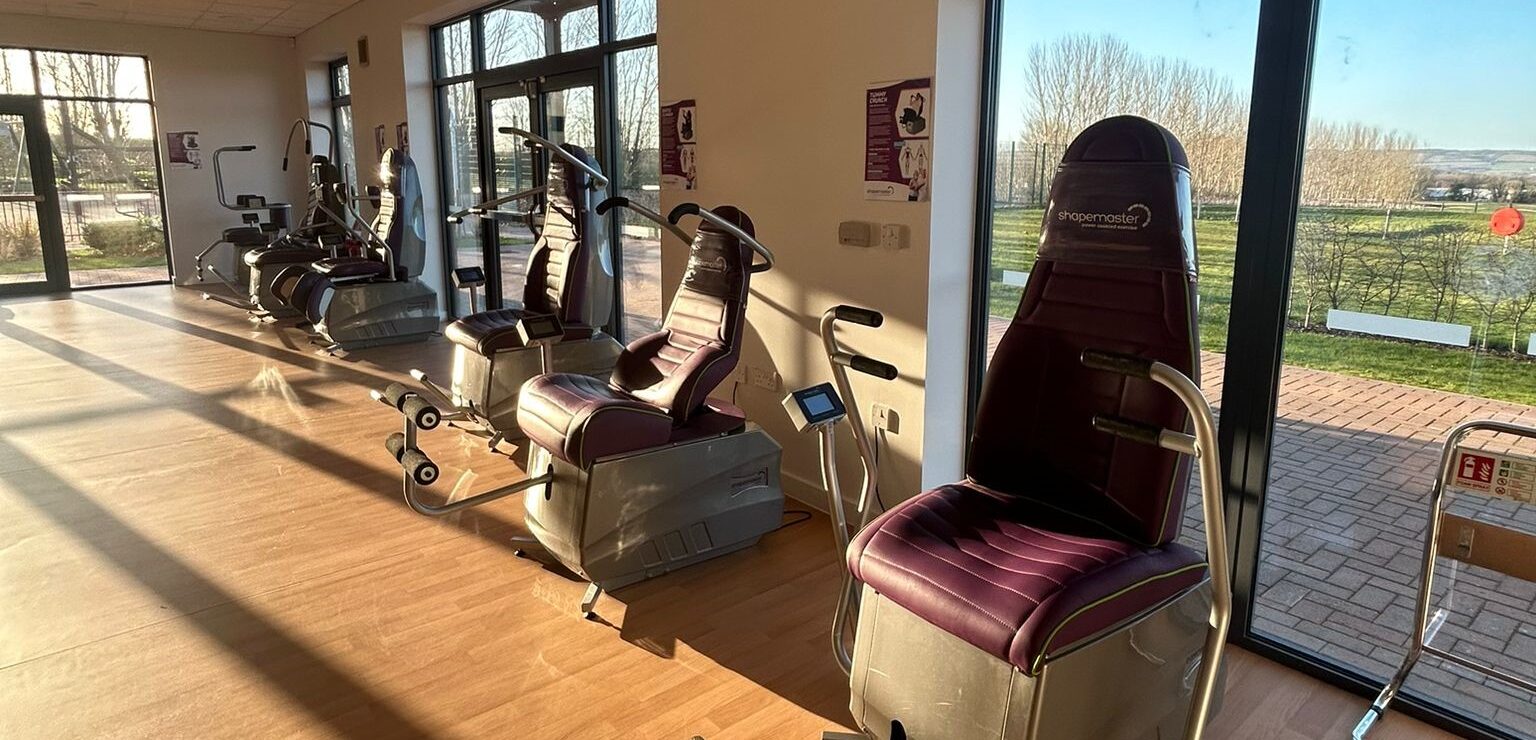 A row of Shapemaster power-assisted exercise machines in a bright KIMS Hospital gym with large windows and garden views.