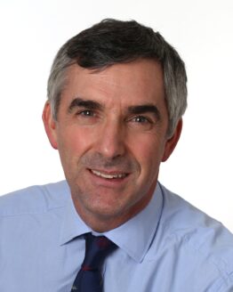 Headshot of a man with salt-and-pepper hair, wearing a light blue dress shirt and a dark tie on a grey background.