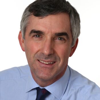 Headshot of a man with salt-and-pepper hair, wearing a light blue dress shirt and a dark tie on a grey background.