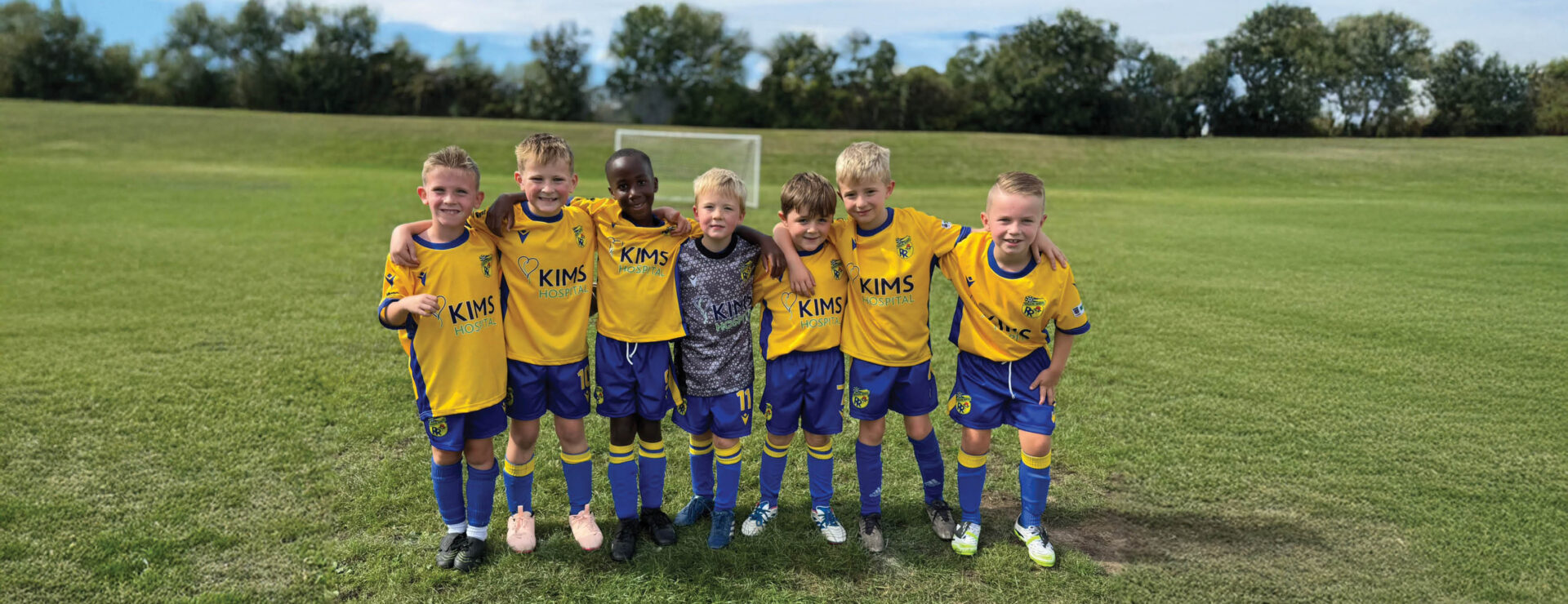 A youth football team in yellow KIMS Hospital sponsored kits stand with arms around each other on a grass pitch.