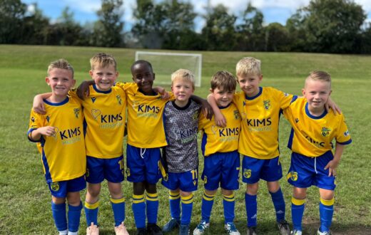 A youth football team in yellow and blue KIMS Hospital kits standing together on a grass pitch outdoors.