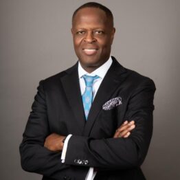 Professional portrait of a smiling man with crossed arms, wearing a black suit, light blue tie, and pocket square.