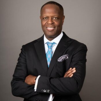 Professional portrait of a smiling man with crossed arms, wearing a black suit, light blue tie, and pocket square.