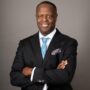 Professional portrait of a smiling man with crossed arms, wearing a black suit, light blue tie, and pocket square.