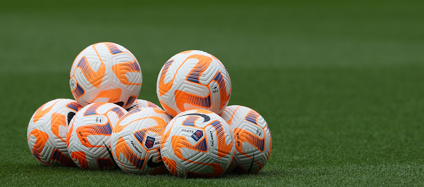 A stack of white and orange Nike Flight footballs sitting on a green grass pitch, ready for a match or training.